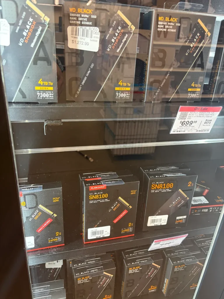 Boxes of WD Black SN850X and SN8100 NVMe SSDs are displayed on a store shelf, showing capacities such as '4TB' and '2TB' and performance details like '7300MB/s'; a price tag reads '$1,272.99'.