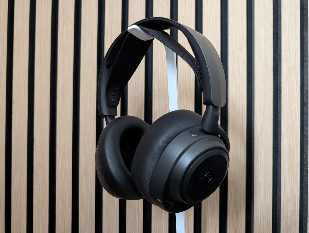 A pair of SteelSeries Arctis Nova Pro headphones hangs in front of a wooden slatted wall.