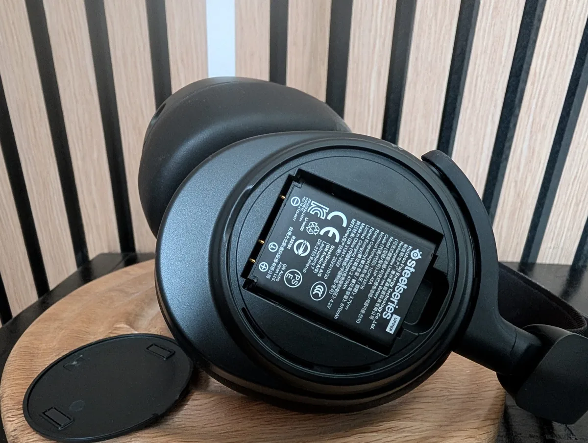 A SteelSeries Arctis headset with the battery compartment open, showing the battery labeled'SteelSeries.'