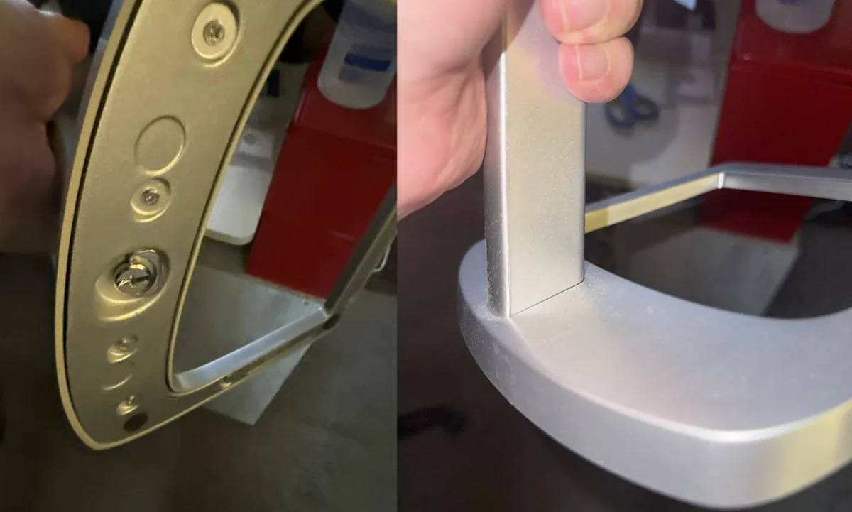 Close-up of a hand holding a metallic U-shaped handle or frame, showing screws and rounded edges, photographed indoors with a workbench and tools blurred in the background