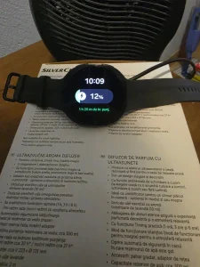 Galaxy Watch 9 carga lenta problema — Black Samsung Galaxy Watch 9 resting in front of a desk fan showing 12 percent battery charge at 10:09 AM at the start of a cooling fast charge test.