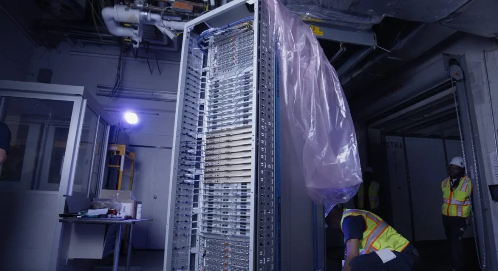 A large, vertically standing, unbranded server rack filled with equipment is being installed by workers in a dimly lit data center.