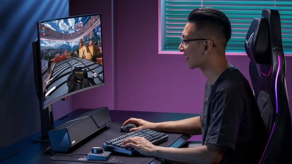 A man plays a game on his PC while the ROG Gjallar gaming soundbar sits on his black desk surrounded by ROG gear.