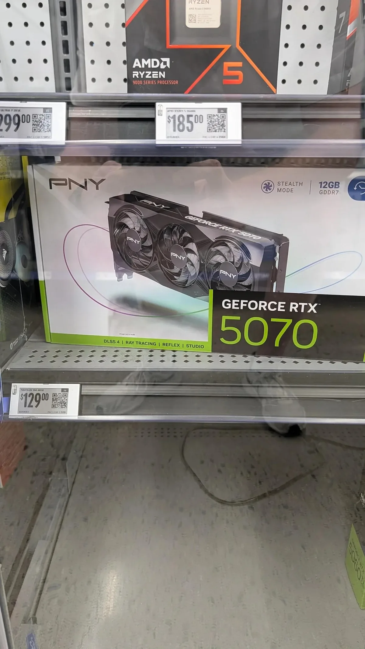 RTX 5070 precio Walmart error — A PNY GeForce RTX 5070 graphics card box is displayed on a shelf next to an AMD Ryzen 5 9000 Series processor, with the card priced at $129 and the processor at $185.