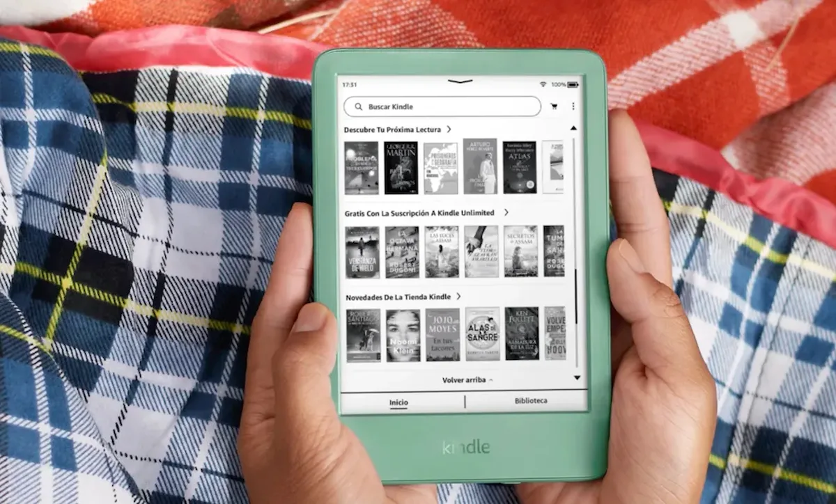 Amazon kindle — Manos sosteniendo un lector electrónico Kindle en color verde claro mostrando el catálogo de libros sobre una manta de cuadros.