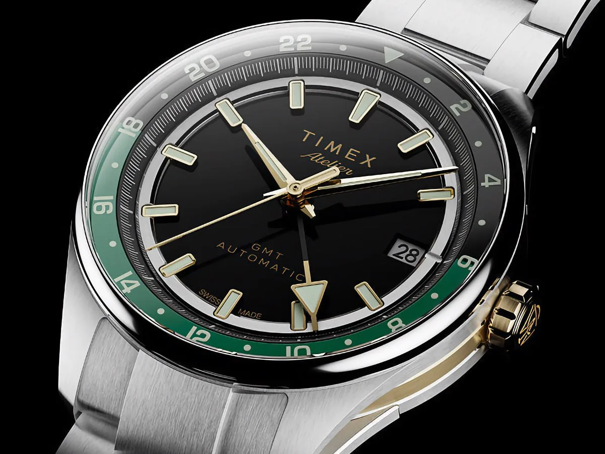 Angled view of the Timex Atelier GMT Première M1a showing its black dial, green GMT ring, gold-tone crown, and stainless steel bracelet.