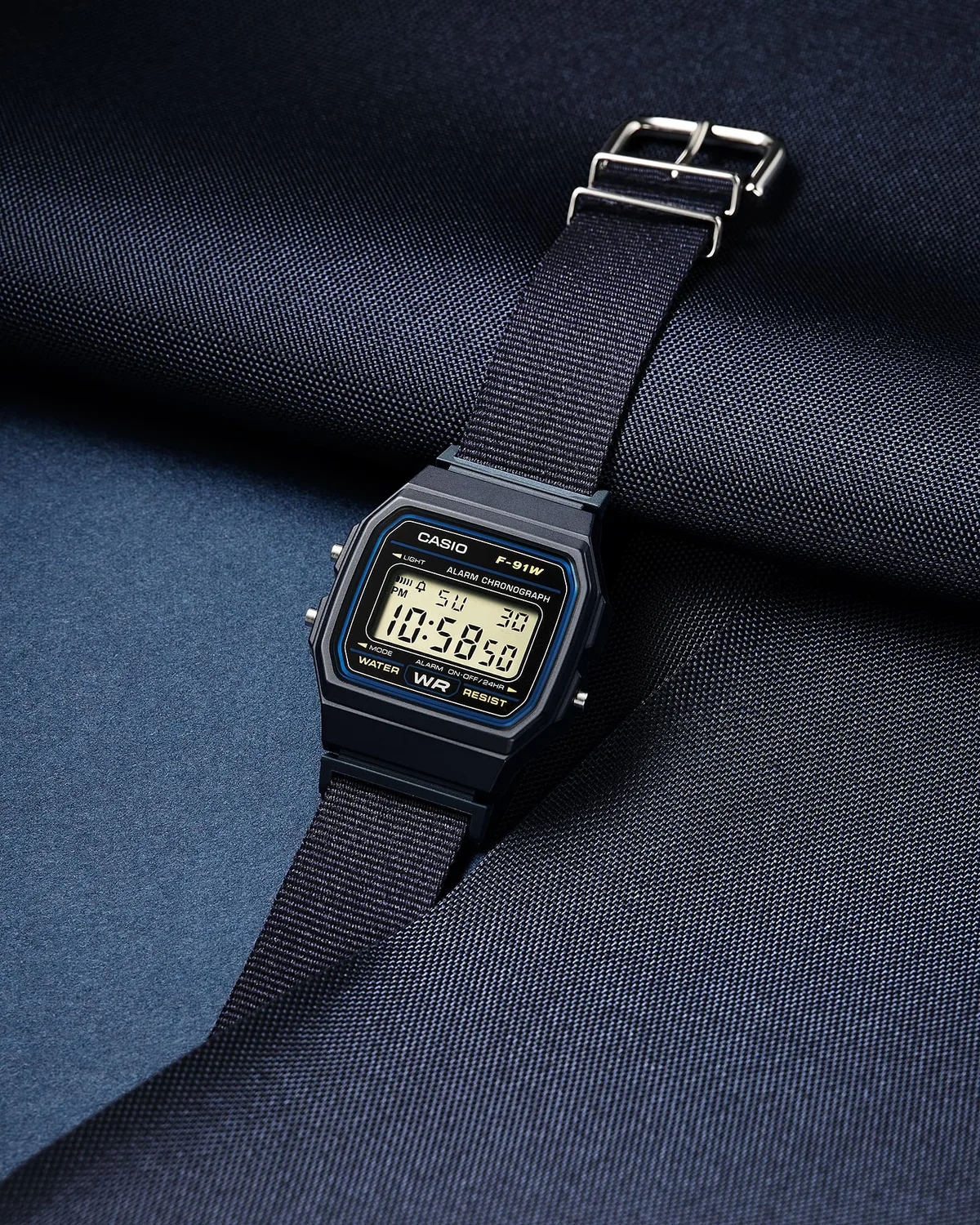 casio f 91w correa — Casio F-91WF-2A digital watch with a black case, navy blue woven fabric strap, and silver accents, photographed against a navy blue textured background.