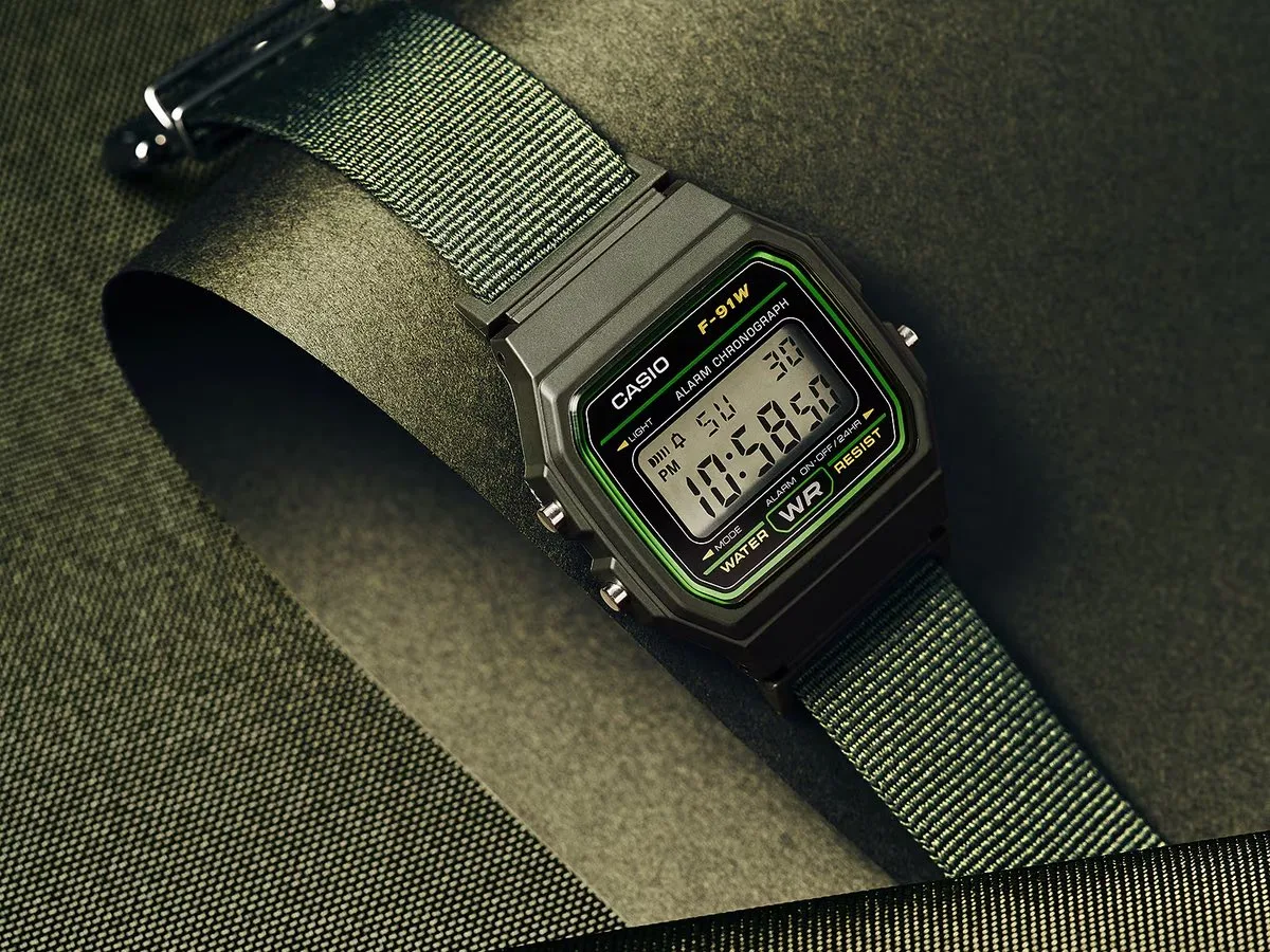 Casio F-91WF-3A digital watch with a green-accented black case and woven green fabric strap, photographed against a dark green textured background.