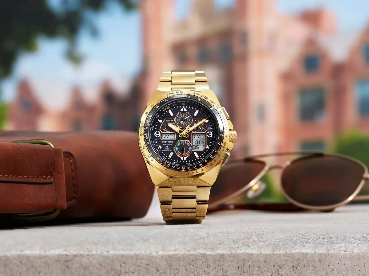 citizen promaster skyhawk titanium — Citizen Promaster Skyhawk A-T JY8144-50E gold-tone pilot watch displayed upright on a ledge beside a brown leather bag and sunglasses, with a blurred cityscape in the background.