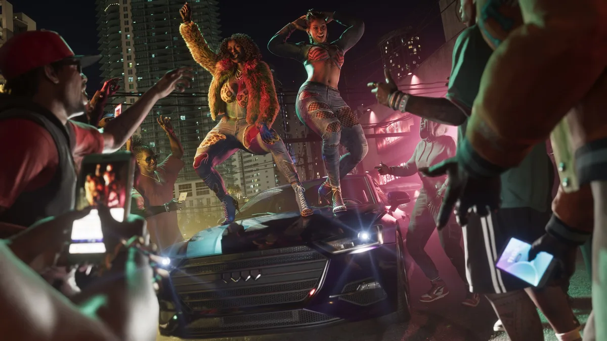 Two characters dance on a car in a lively street scene from the game “Grand Theft Auto VI,” surrounded by cheering people using smartphones.