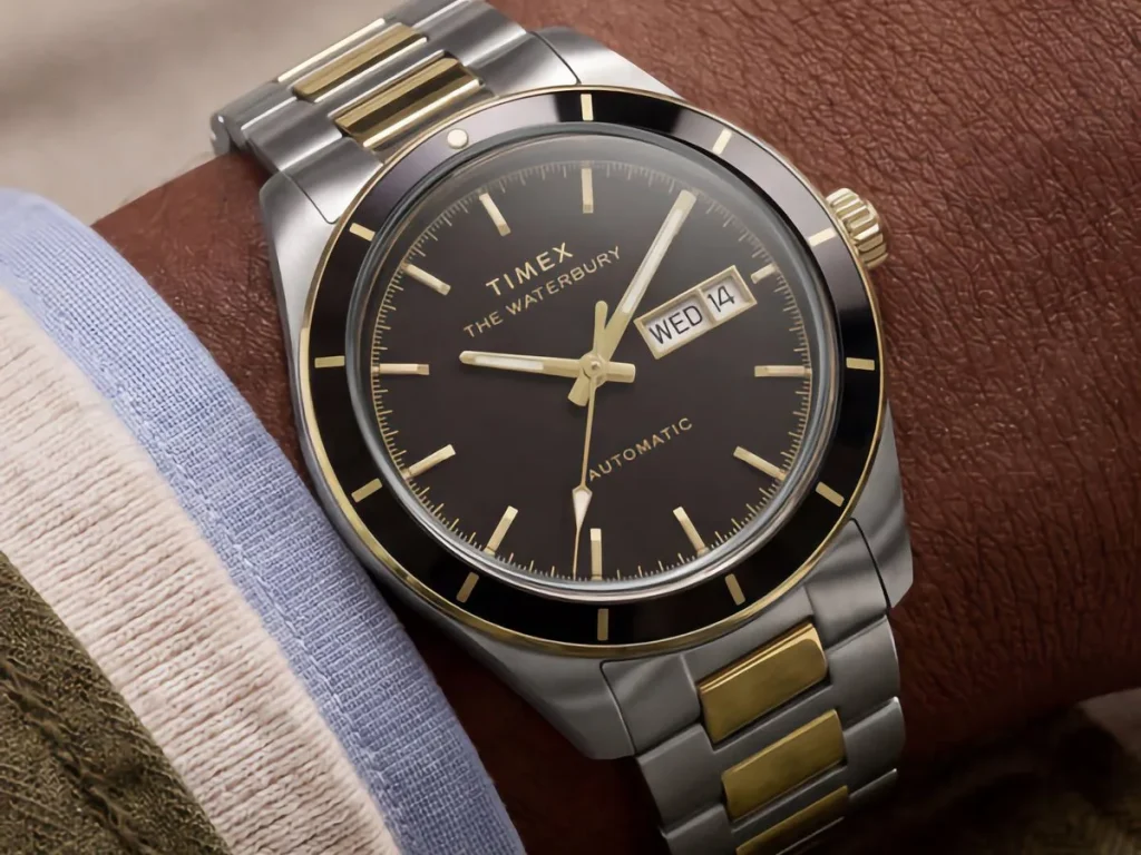 reloj timex waterbury heritage — Two-tone Timex Waterbury Heritage Automatic with black dial worn on a wrist, showing its day-date display and gold-tone accents.
