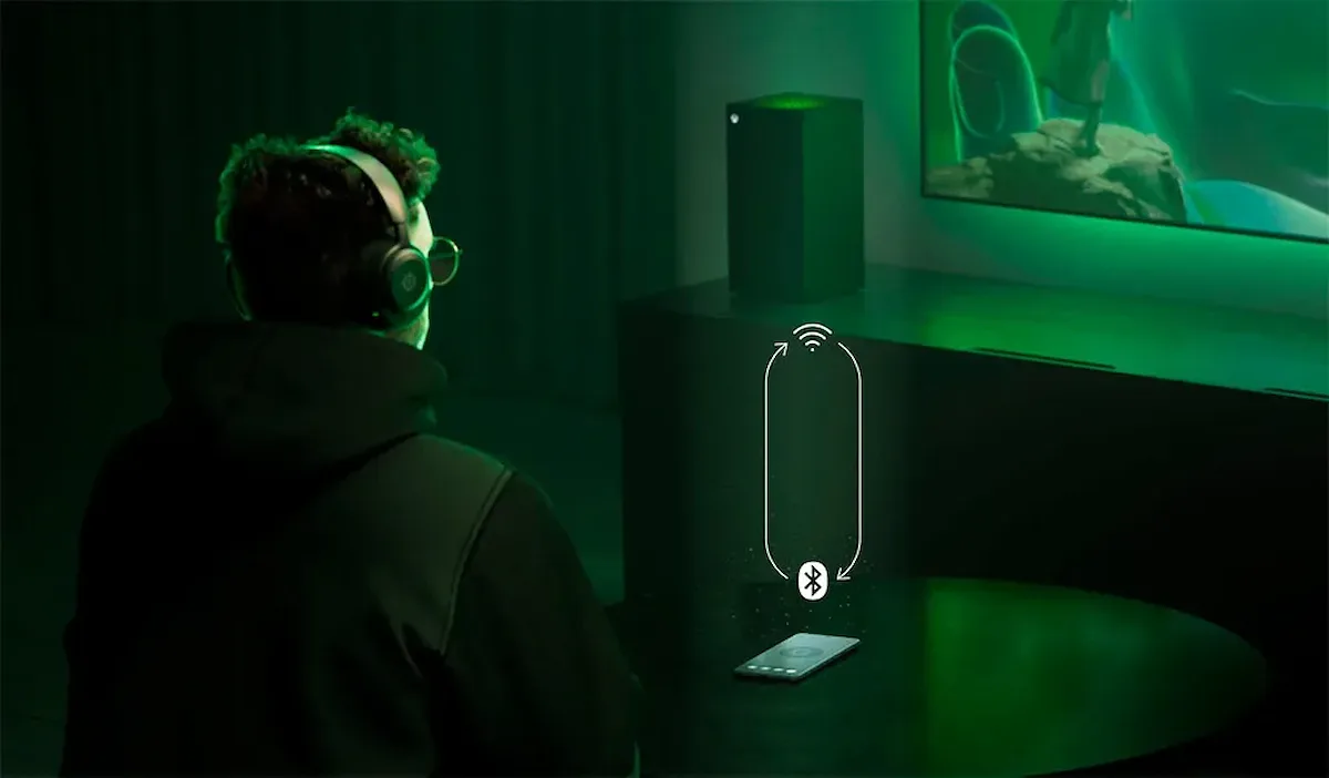 Usuario con auriculares SteelSeries Arctis Nova 5X frente a una consola y un smartphone, ilustrando la conectividad inalámbrica dual.