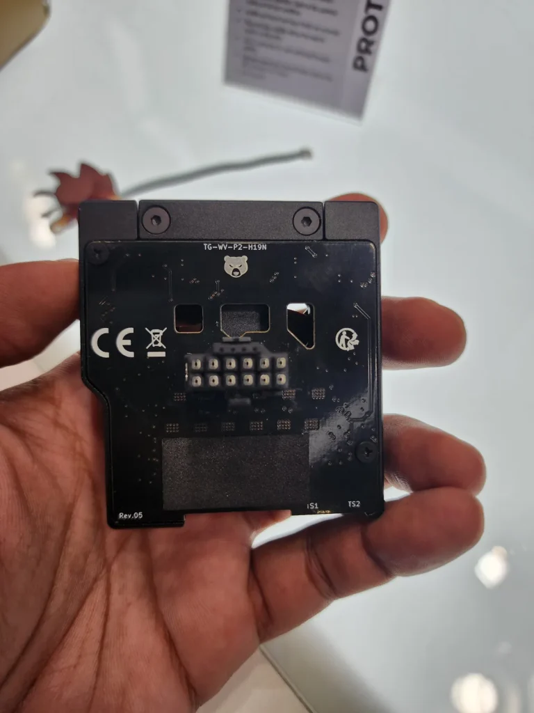 A person holds a component labeled 'TG-WV-P2-H19N' and 'Rev.05', with visible connectors and a partially obscured display stand in the background.