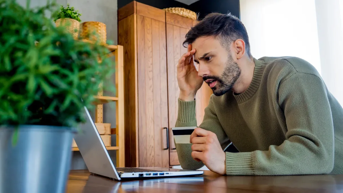 Hombre preocupado con un portátil y una tarjeta de crédito en una cocina, ante la crisis de la RAM y portátiles más caros