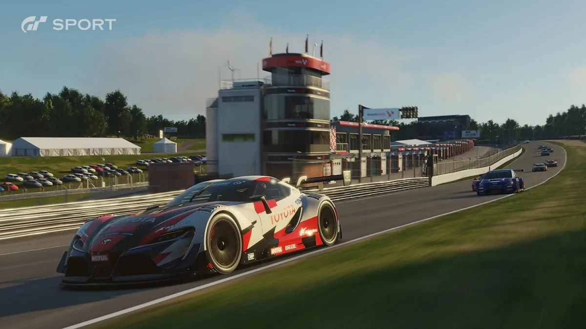 Coche de carreras Toyota toma una curva en Gran Turismo Sport en PC, con otros coches detrás y el edificio de boxes junto al circuito