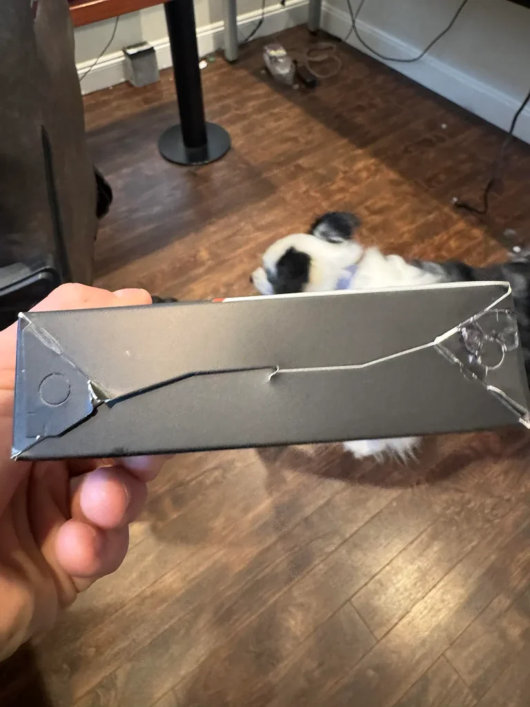 A hand holding a damaged black cardboard box with cracks and tears, while a dog is visible in the background on a wooden floor.