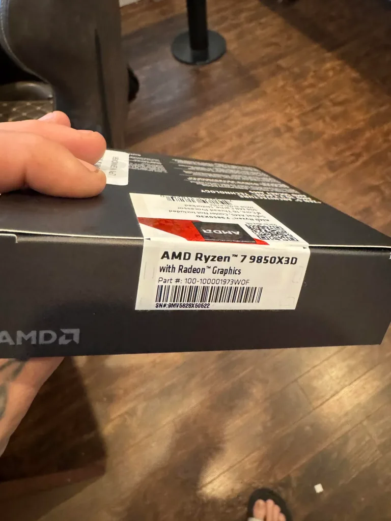 Intel Core — A person holding a box labeled AMD Ryzen 7 9850X3D with Radeon Graphics in a room with hardwood flooring.
